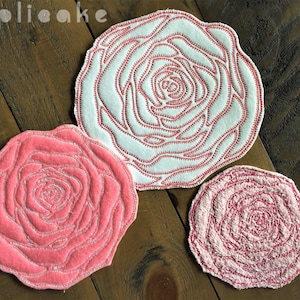 May include: Three embroidered rose-shaped fabric coasters in different sizes. The largest coaster is white with a pink outline, the medium coaster is pink with a white outline, and the smallest coaster is light pink with a darker pink outline.