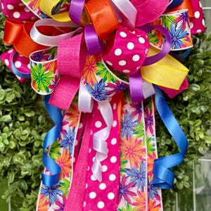 Puede incluir: Un lazo decorativo hecho de múltiples cintas en colores vibrantes que incluyen rosa fuerte, naranja, amarillo, morado y azul. Las cintas presentan patrones de lunares blancos e impresiones florales multicolores, con largas colas que cuelgan hacia abajo.