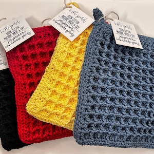 Puede incluir: Cuatro posavasos de ganchillo en negro, rojo, amarillo y azul. Cada posavasos tiene una etiqueta que dice "hot pad" e incluye detalles sobre el material, las instrucciones de cuidado y el nombre del fabricante, "jellybeeaaan".