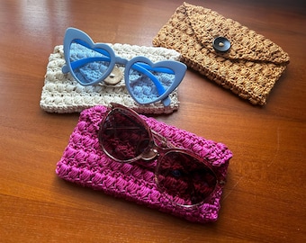 May include: Three pairs of sunglasses rest on crocheted cases. A pair of blue heart-shaped sunglasses sits on a cream-colored case. A pair of brown sunglasses rests on a fuchsia case. A tan case with a black button closure is also present.