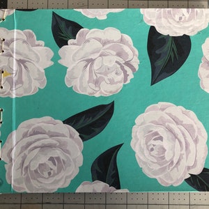May include: A handmade journal with a floral design. The cover features large white roses with gray shading and dark green leaves on a turquoise background. The binding is hand-stitched with white thread.