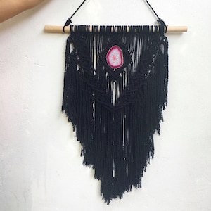 May include: Black macrame wall hanging with a pink and white agate stone in the center. The hanging is attached to a wooden dowel rod.