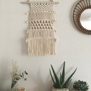 May include: A macrame wall hanging with a natural cream color and a wooden dowel at the top. The hanging has a fringe at the bottom and is made with a variety of knots. There are three potted plants in the foreground, two with macrame plant hangers.
