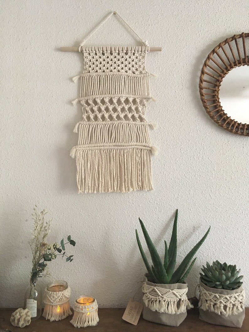 May include: A macrame wall hanging with a natural cream color and a wooden dowel at the top. The hanging has a fringe at the bottom and is made with a variety of knots. There are three potted plants in the foreground, two with macrame plant hangers.