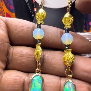 May include: Gold dangle earrings with a green opal stone, gold fish charms, and white beads.