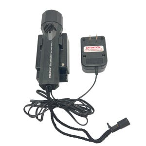 May include: A black Pelican StealthLite rechargeable flashlight with a charging base and a wall plug adapter. The flashlight body features the text Pelican StealthLite Rechargeable. The wall adapter has a label reading ATTENTION: CHARGE 24 HOURS BEFORE USE. TURN LIGHT OFF WHILE CHARGING.