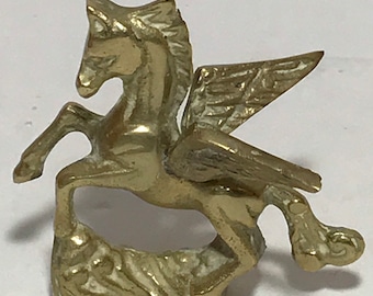 Puede incluir: Una figurita de bronce de color dorado de un caballo alado, o pegaso, con las alas extendidas. El caballo está de pie sobre una base rocosa.