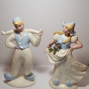 May include: A pair of vintage ceramic figurines depicting a boy and girl in traditional Dutch clothing. The boy is wearing a blue hat and vest, white pants, and blue shoes. The girl is wearing a blue and white dress with white polka dots, a yellow hat, and blue shoes. Both figurines are standing on a white base.