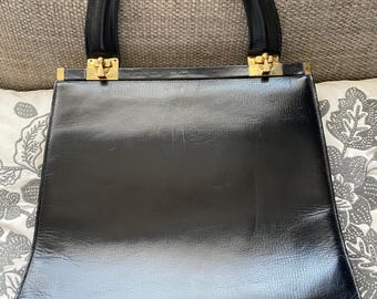 Puede incluir: Un bolso de mano de cuero negro con herrajes dorados y dos asas superiores.