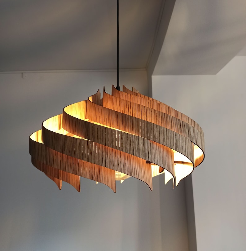 Może przedstawiać: Nowoczesna, drewniana lampa wisząca o unikalnym, warstwowym designie. Lampa wykonana jest z cienkich, zakrzywionych pask&oacute;w drewna, kt&oacute;re są ułożone jedna na drugiej, tworząc efekt spirali. Lampa jest oświetlona od wewnątrz, dając ciepłe światło.