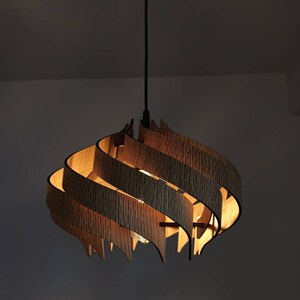 May include: A wooden pendant light fitting with a unique, swirling design. The light is made of multiple layers of wood, creating a layered effect. The light is on and casting a warm glow.