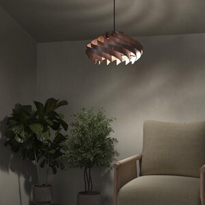 May include: A modern, wooden pendant light fixture with a spiral design. The light is suspended from a black cord and ceiling mount. The fixture is made of layered, curved wooden slats, casting warm light. The room features plants and a neutral-toned armchair.