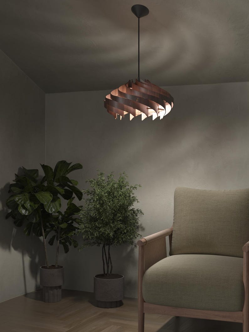 May include: A modern, wooden pendant light fixture with a spiral design. The light is suspended from a black cord and ceiling mount. The fixture is made of layered, curved wooden slats, casting warm light. The room features plants and a neutral-toned armchair.
