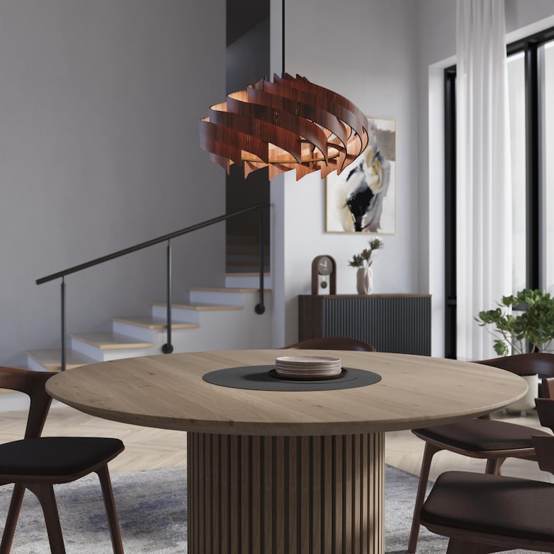 May include: A modern dining room featuring a large, round wooden table with a dark center and a ribbed base. Above the table hangs a decorative, layered wooden pendant light. The room includes chairs, a staircase, and artwork.