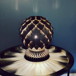 Op de afbeelding: Een houten lamp in de vorm van een dennenappel met een licht dat door het ingewikkelde ontwerp schijnt. De lamp staat op een zwarte tafel.