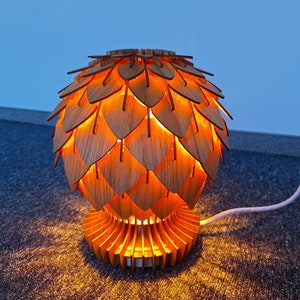 May include: A wooden table lamp with a unique design. The lamp features a layered, petal-like shade and a base with a fan-like structure. The warm light from the bulb creates a cozy ambiance. The lamp is powered by a white cord.