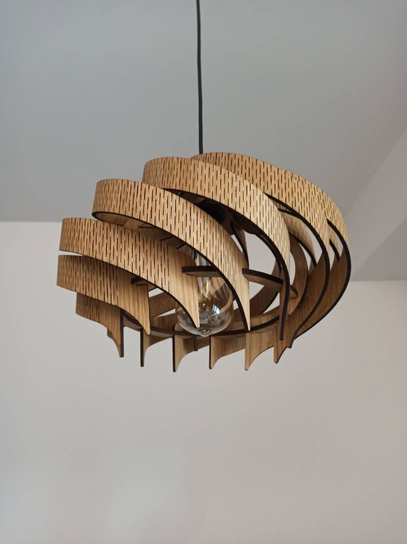 May include: A wooden pendant light fitting with a unique, geometric design. The light fitting is made of multiple layers of wood that are stacked and curved to create a spiral shape. The light fitting is suspended from the ceiling by a black cord.