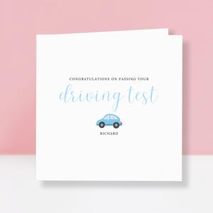 May include: A white card with the text "CONGRATULATIONS ON PASSING YOUR driving test" in light blue script. A small blue car graphic is centered below the text, with the name "RICHARD" below the car. The card is on a pink background.