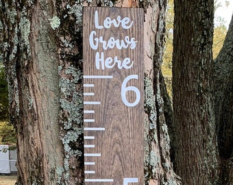 May include: A wooden growth chart with white numbers and the text "Love Grows Here" on a brown wooden background. The chart is mounted on a tree trunk.