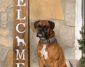 May include: A wooden sign with the word "WELCOME" spelled vertically in white letters. A white silhouette of a dog is between the letters "C" and "M". A brown and white boxer dog is sitting in front of the sign.