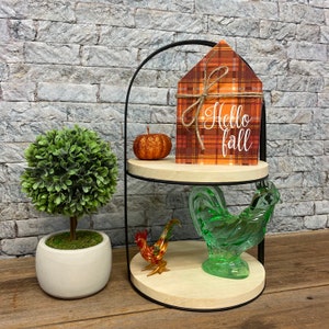 May include: A two-tiered black metal stand with a wooden top. The top tier features a plaid house-shaped sign with the text "Hello Fall" and a small orange pumpkin. The bottom tier has a green glass rooster figurine and a small red and orange rooster figurine.