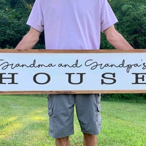 May include: A white wooden sign with brown trim, featuring the text "Grandma and Grandpa's HOUSE" in black lettering. The sign is being held by a person.