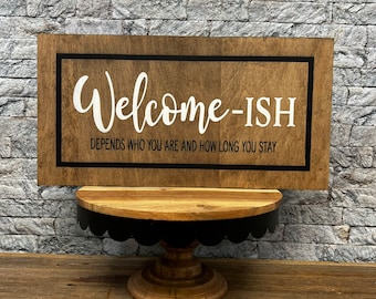 May include: Wooden sign with the text "Welcome-ish" and the caption "Depends who you are and how long you stay". The sign is on a black and brown cake stand.
