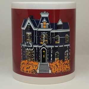 May include: A red ceramic mug with a white rim features a black and white illustration of a spooky house with boarded up windows and overgrown vines. The house is set against a backdrop of orange bushes.