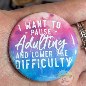 May include: A round button with a watercolor background in shades of pink, blue, and purple. The button features the text "I WANT TO - PAUSE - Adulting AND LOWER THE DIFFICULTY" in white script. The button is held in a hand.