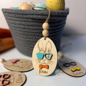 May include: Wooden Easter egg decorations featuring engraved bunny faces, each with a different coloured bow tie and glasses. One ornament says "Beau". A grey basket filled with Easter eggs is in the background.