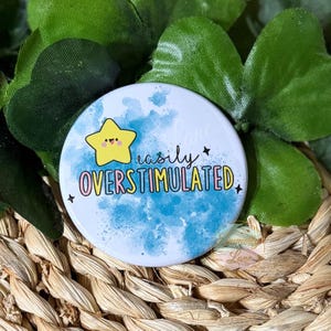 May include: A round, white button with the text "easily OVERSTIMULATED" in a colorful font. A yellow star with a smiling face is on the left. The background has a blue watercolor effect. The button is surrounded by green leaves and woven basket.