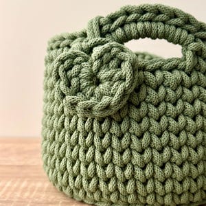 May include: A sage green crochet basket crafted from thick cord, featuring a rounded body with a textured stitch pattern. The basket includes a sturdy integrated handle and a decorative circular crochet flower accent attached to the front side.