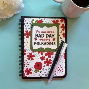 May include: A spiral-bound notebook featuring a white cover with a red floral pattern and a central green label. The text reads: You can't have a BAD DAY wearing POLKADOTS, accompanied by two small ladybug illustrations. A black pen and a white pen rest on top.