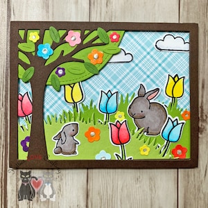 May include: A brown card with a die-cut window showing a green tree with pink, blue, and yellow flowers. Two gray bunnies are in the grass with pink, blue, and yellow tulips.