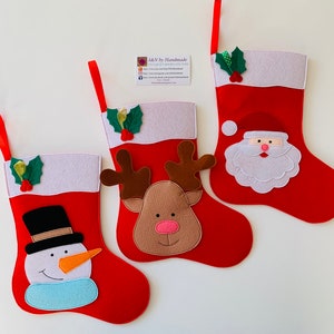 May include: Four felt Christmas stockings featuring different festive characters. The stockings are red with white cuffs and have a green holly sprig at the top. The characters include a snowman, a reindeer, Santa Claus, and a generic Christmas character.