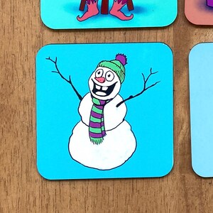 May include: A blue square coaster with a cartoon snowman wearing a green and purple scarf and a green hat with a purple pom-pom. The snowman has a big smile and is standing with its arms outstretched.