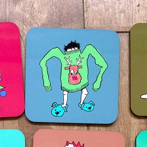 May include: A blue square coaster with a cartoon illustration of a person wearing a green sweater with a reindeer design and blue slippers. The sweater has the text "Merry Christmas" on it.