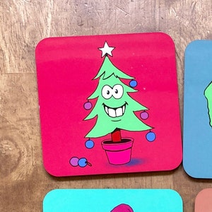May include: A red square coaster with a green Christmas tree with a smiling face and a pink pot. The tree has blue and pink ornaments.