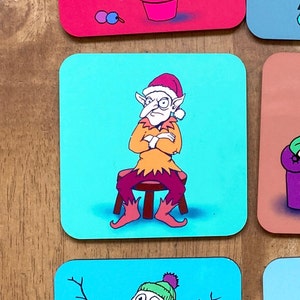 May include: A blue and white coaster with a cartoon illustration of a grumpy elf wearing a red hat and orange and yellow clothing. The elf is sitting on a brown stool with his arms crossed.