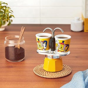 May include: A yellow Mini Express stovetop espresso maker with two cups, each with a colourful design. The cups are filled with coffee. A glass jar of coffee grounds and a wooden spoon are on the left.