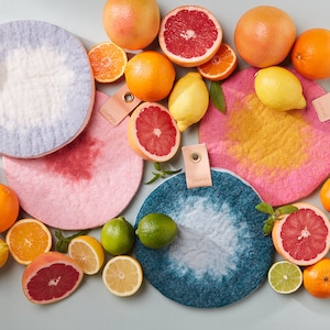May include: A selection of colourful felt trivets in blue, pink, and yellow, displayed with fresh citrus fruits including grapefruits, oranges, lemons, and limes. Each trivet has a leather tag with the word "aveva".