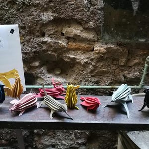 May include: A collection of colorful, wooden animal sculptures, including an owl, rooster, and various other creatures. The sculptures are arranged on a dark surface, with a white card featuring the "lovi" logo in the background. The animals are in shades of blue, pink, yellow, red, and black.