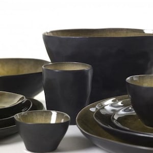 Puede incluir: Colección de vajilla de cerámica negra con interior dorado en contraste. El juego incluye cuencos, platos y tazas de varios tamaños. Las piezas tienen un acabado mate y una forma ligeramente irregular, lo que les da un aspecto artesanal.