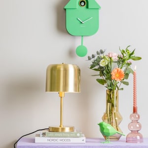 May include: A green cuckoo clock with a pendulum hanging from a white wall. A gold table lamp with a white lampshade sits on a purple table with a book titled "Nordic Moods" and a green origami bird figurine. A pink glass candle holder with a pink candle sits on the table next to a vase with pink and white flowers.