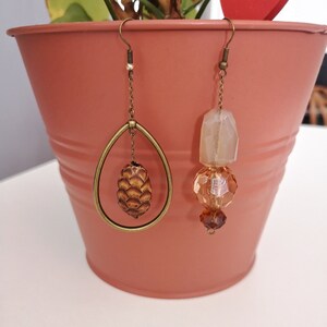 Peut inclure: Une paire de boucles d'oreilles uniques. L'une présente un cadre en forme de goutte d'eau avec un petit pendentif en forme de pomme de pin. L'autre est composée de perles ambrées facettées et rondes, avec une grosse perle de forme irrégulière. Finition dorée.