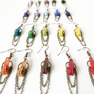 May include: A set of 18 colorful zipper pull earrings with gold chains. The earrings are arranged in a rainbow pattern, with each color represented by a different zipper pull. The zipper pulls are made of plastic and have a shiny finish. The chains are made of metal and have a slightly oxidized finish.