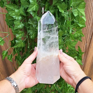 May include: A large, clear quartz crystal point with a natural, raw base. The crystal is held in the hands of a person, showcasing its size and clarity.