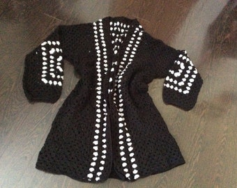 Puede incluir: Un cárdigan de crochet negro con ribetes blancos alrededor de los bordes y en la parte delantera. El cárdigan tiene un frente largo y abierto y mangas largas.