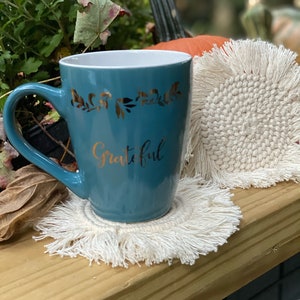 K&ouml;nnte beinhalten: Eine tealblaue Keramiktasse mit goldenem Blumendesign und dem Wort "Grateful" in goldenen Lettern. Die Tasse steht auf einem wei&szlig;en geh&auml;kelten Untersetzer mit Fransen.