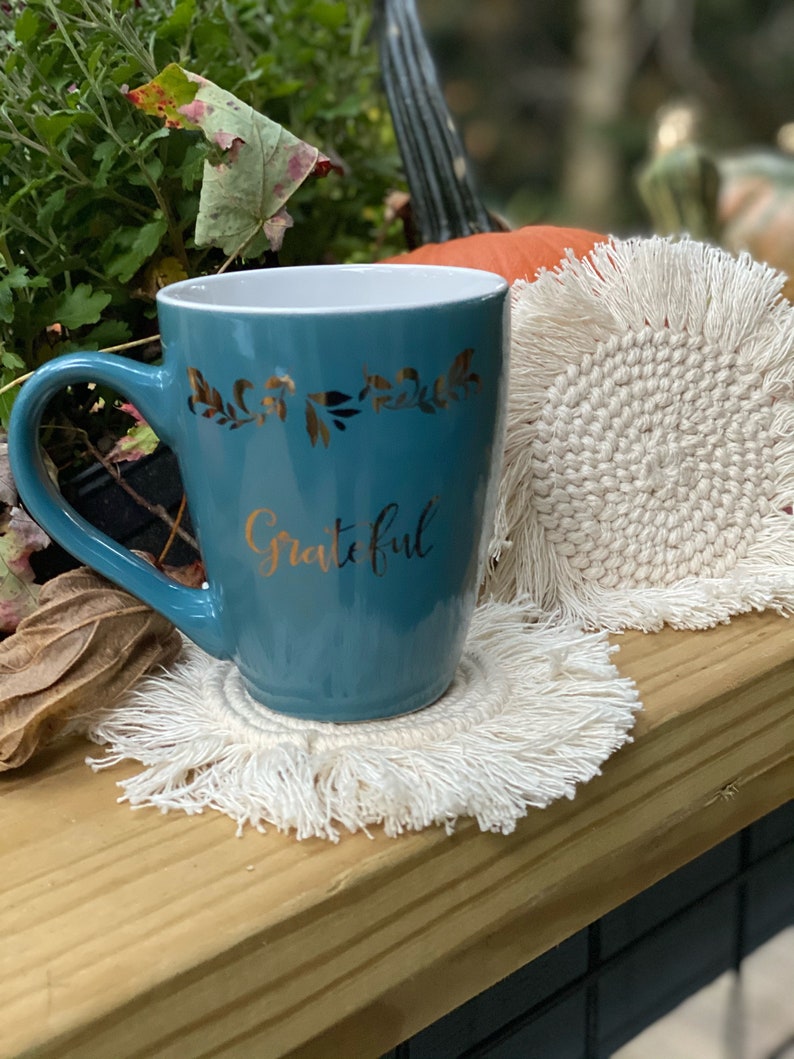 K&ouml;nnte beinhalten: Eine tealblaue Keramiktasse mit goldenem Blumendesign und dem Wort "Grateful" in goldenen Lettern. Die Tasse steht auf einem wei&szlig;en geh&auml;kelten Untersetzer mit Fransen.
